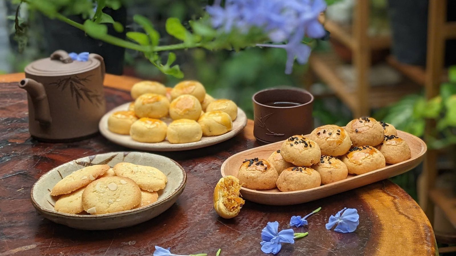 Bàn trà gỗ trong vườn với ba đĩa bánh thủ công của Mộc Mai, ấm trà gốm và những nhành hoa xanh lam
