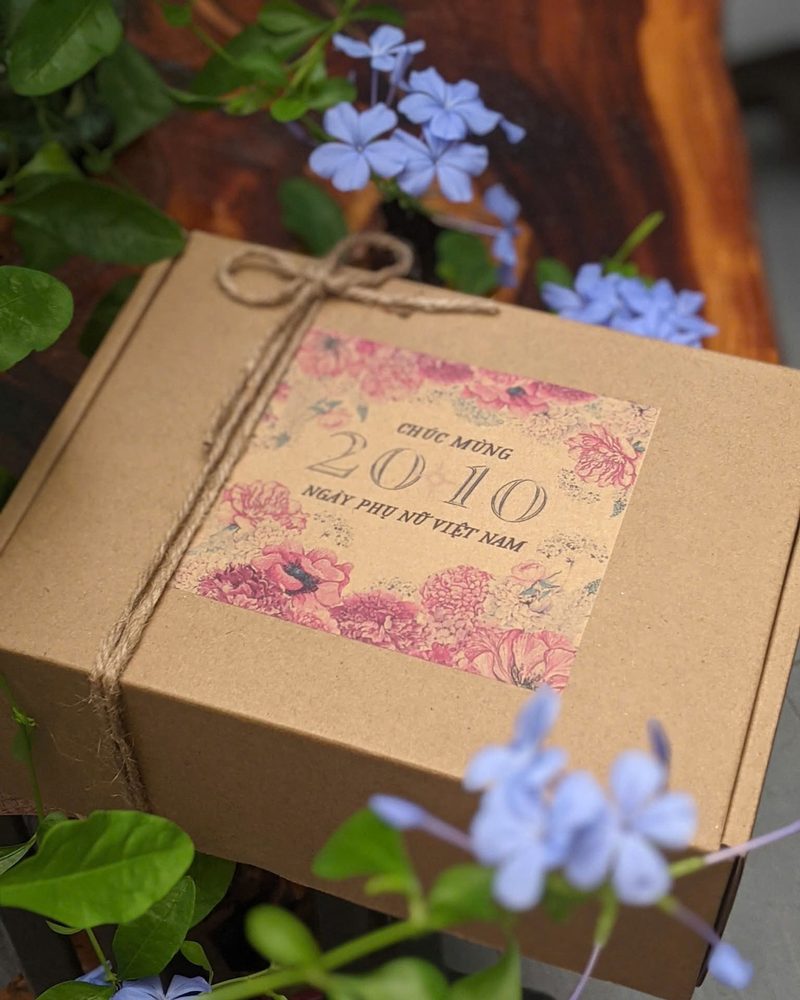Hộp quà kraft buộc dây gai, dán nhãn hoa ‘Chúc mừng 20/10 – Ngày Phụ nữ Việt Nam’, xung quanh là hoa xanh lam và lá non